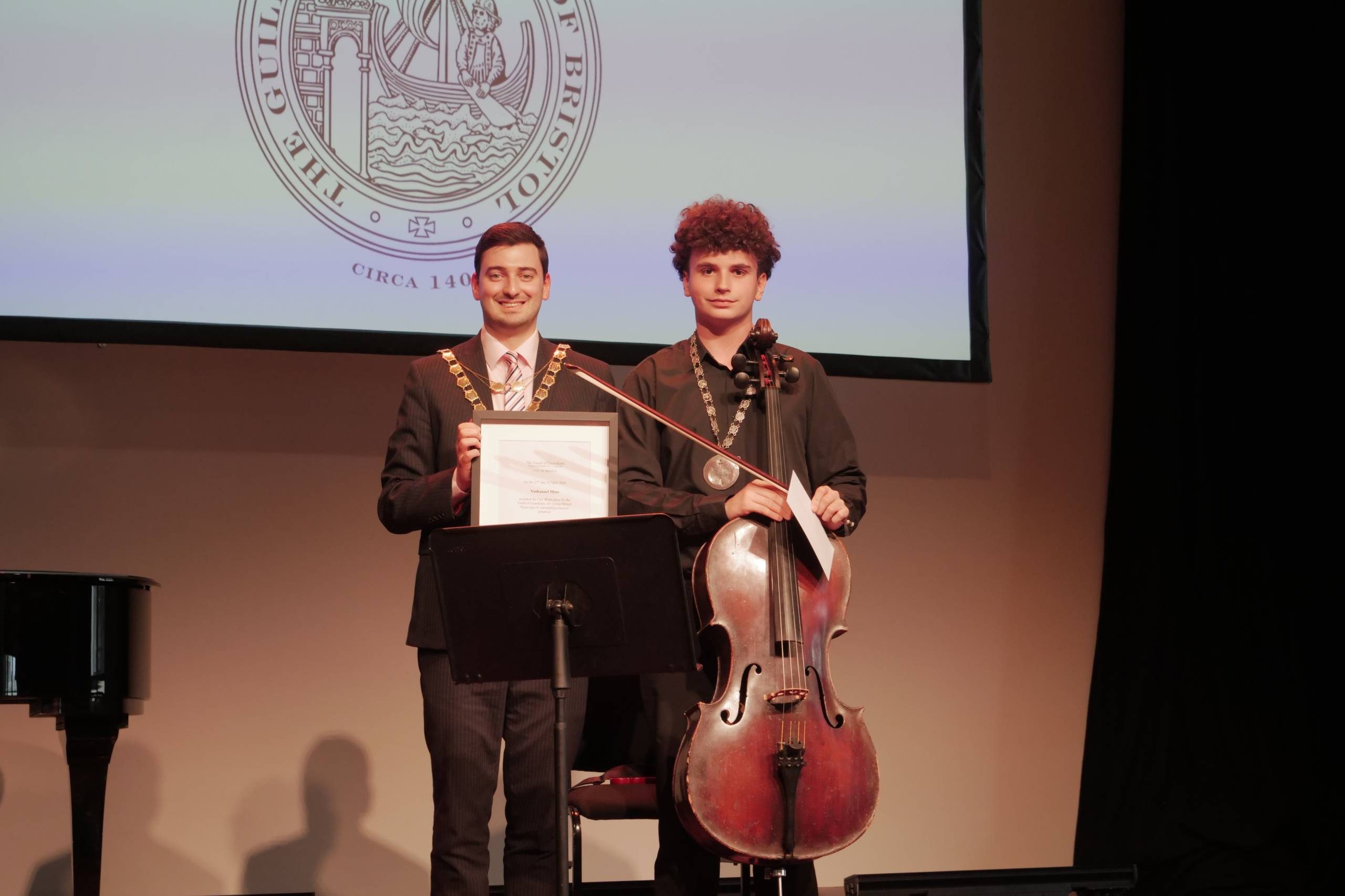 A person wearing a suit and medal holds an award frame, next to a young person holding a cello. They stand on stage