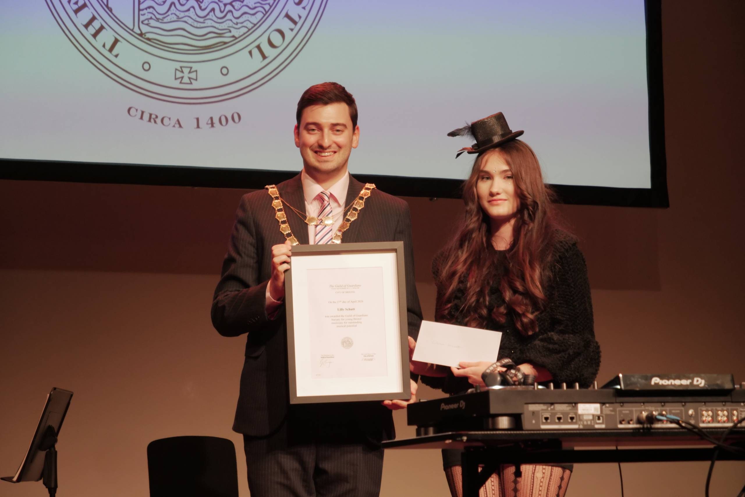 A person wearing a suit and medal holds an award frame, next to a young person stood next to DJ equipment, holding a paper envelope. They stand on stage