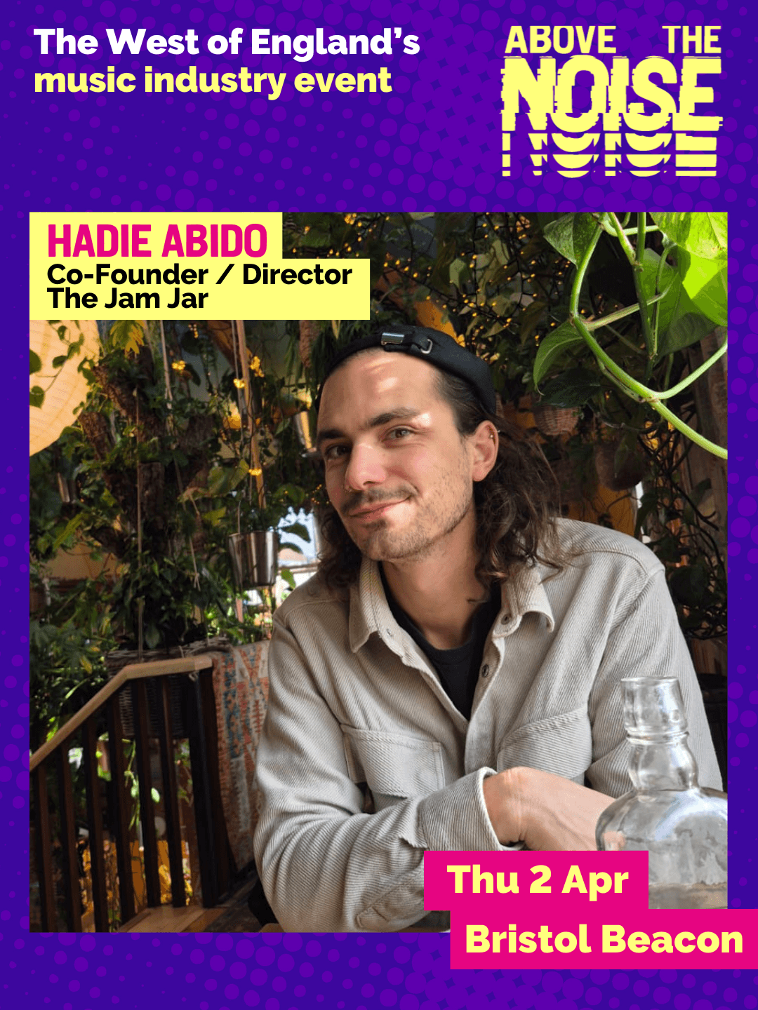 Hadie Abido- Co-Founder / Director, The Jam Jar. Thu 2 Apr, Bristol Beacon. A person with long dark hair wearing an off-white button up shirt poses at a wooden table in front of a wall of lush foliage.