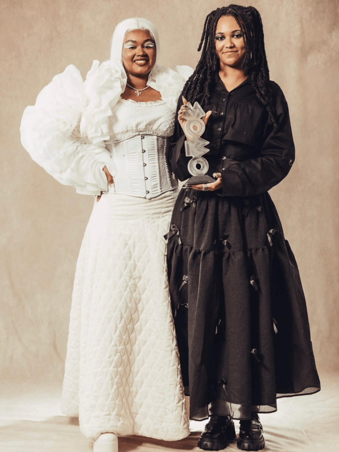 Two people wearing formal attire - one dressed entirely in white, the other in black, pose together, holding an award in the shape of the word 