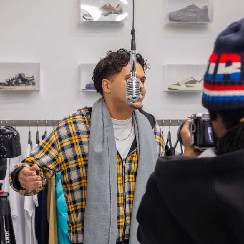 A person wearing a plaid shirt and scarf sings into an old-style microphone hanging in front of them from above.