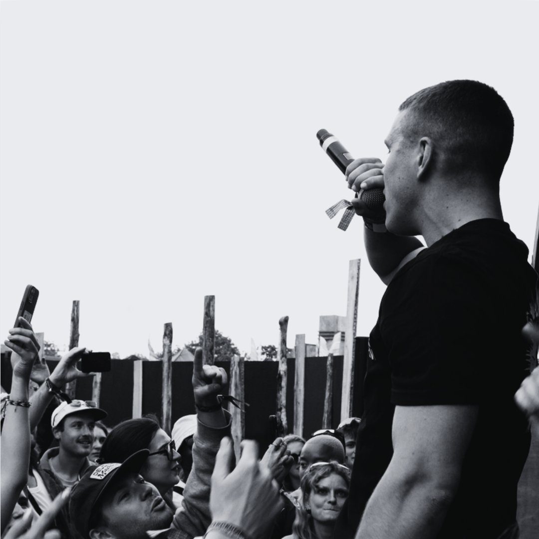 A black and white image of a person wearing a black t-shirt speaking into a microphone in front of a crowd holding their phones to take a video.