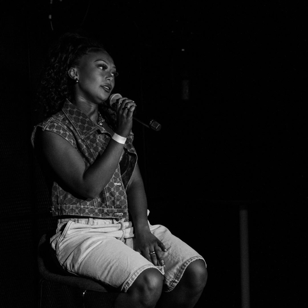 A musician wearing white shorts and jacket sits as they sing into a microphone