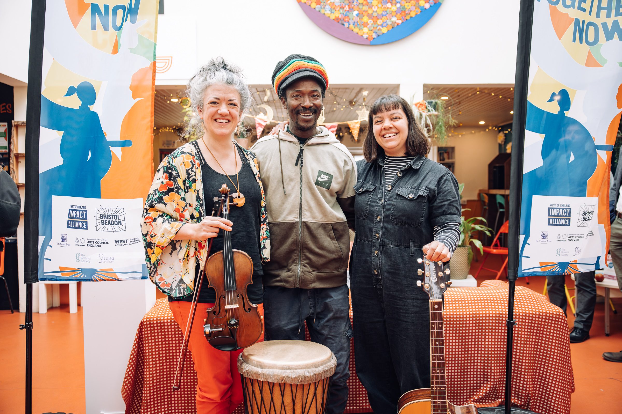Three people stand smiling in a row, in a light foyer space. One holds a violin, there is also a drum and a guitar pictured