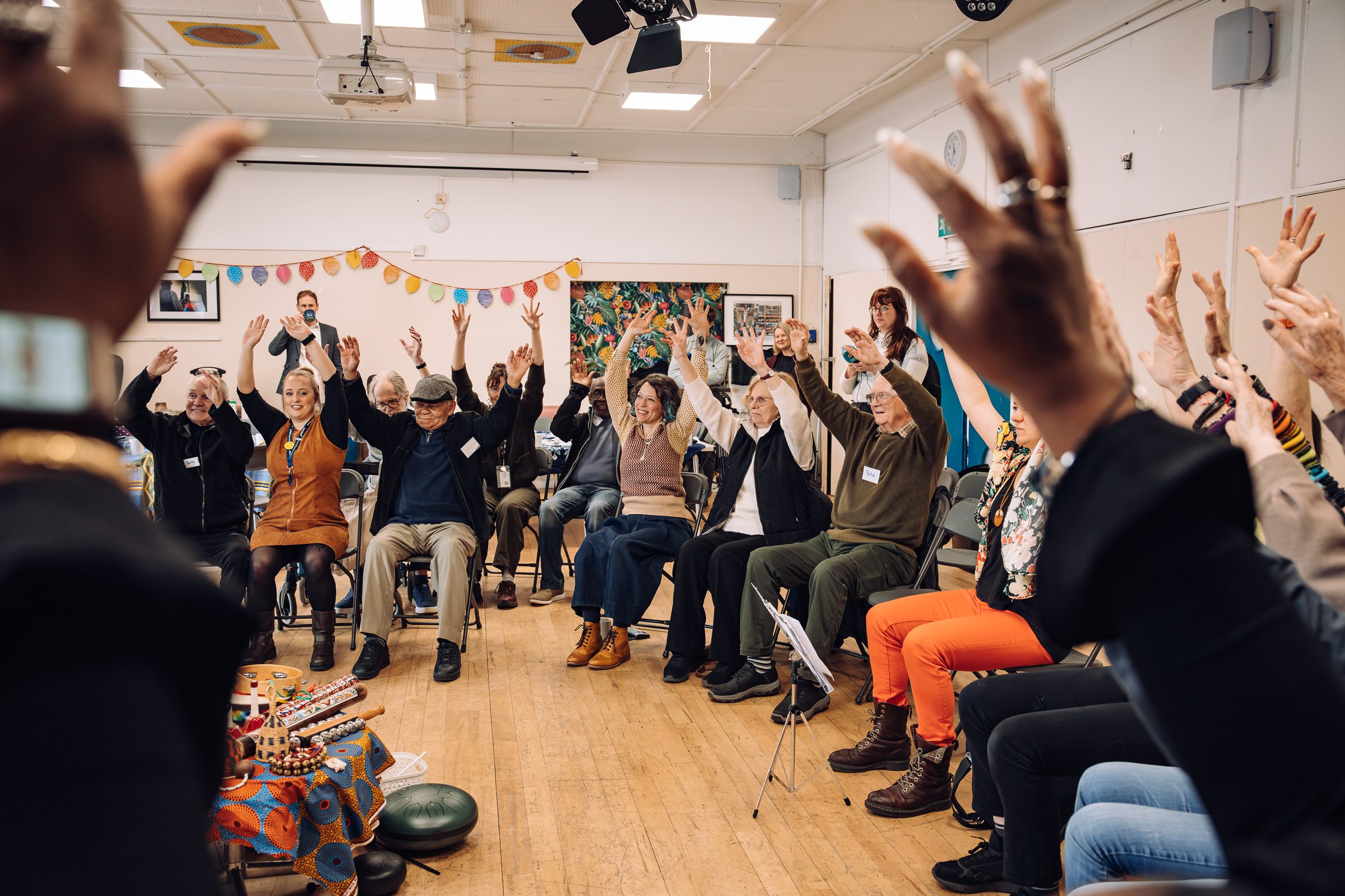 A group of diverse people sit around in a circle with their arms raised joyfully in the air, mid movement