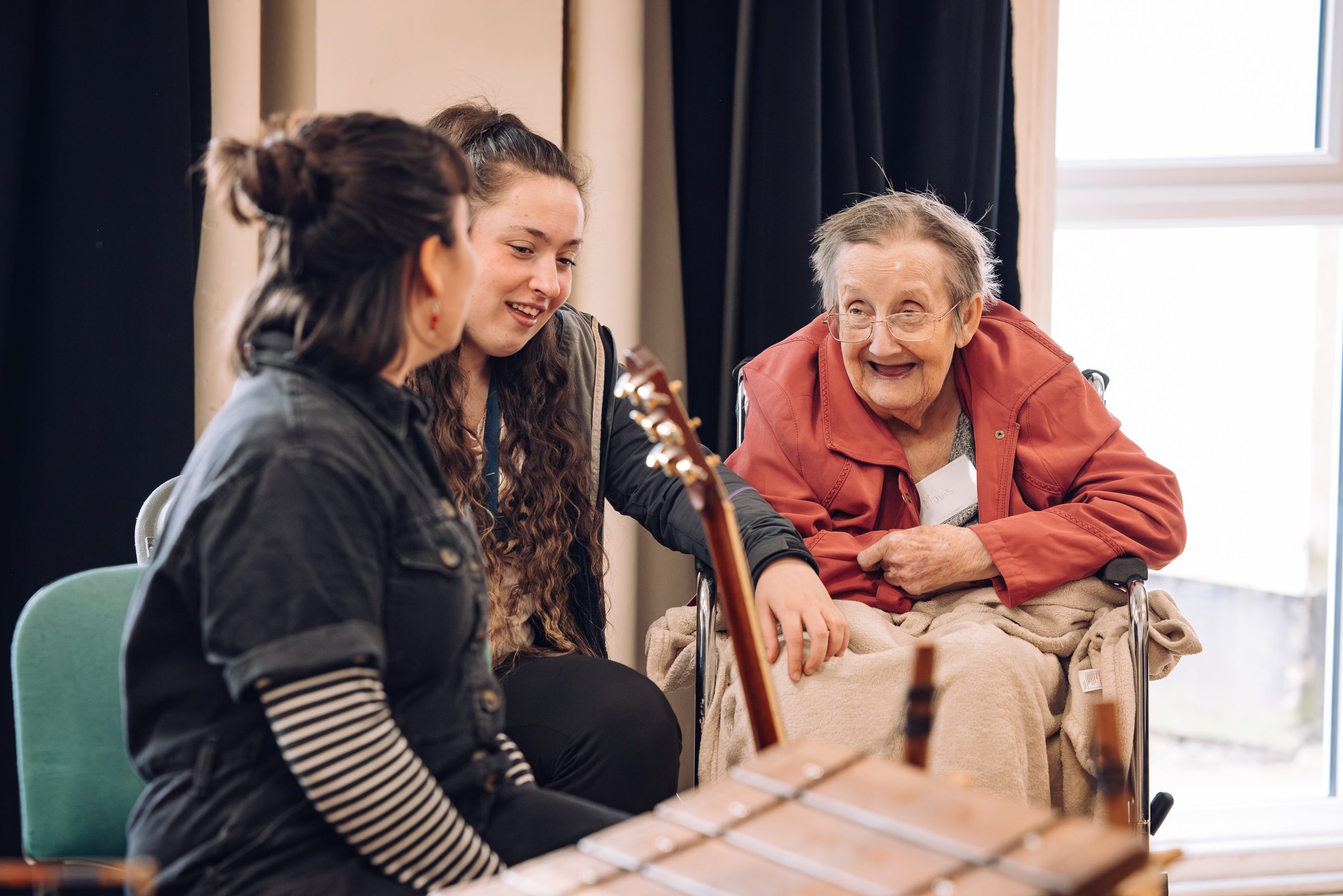 An older person watches and smiles as two younger people, one holding a guitar, sit alongside