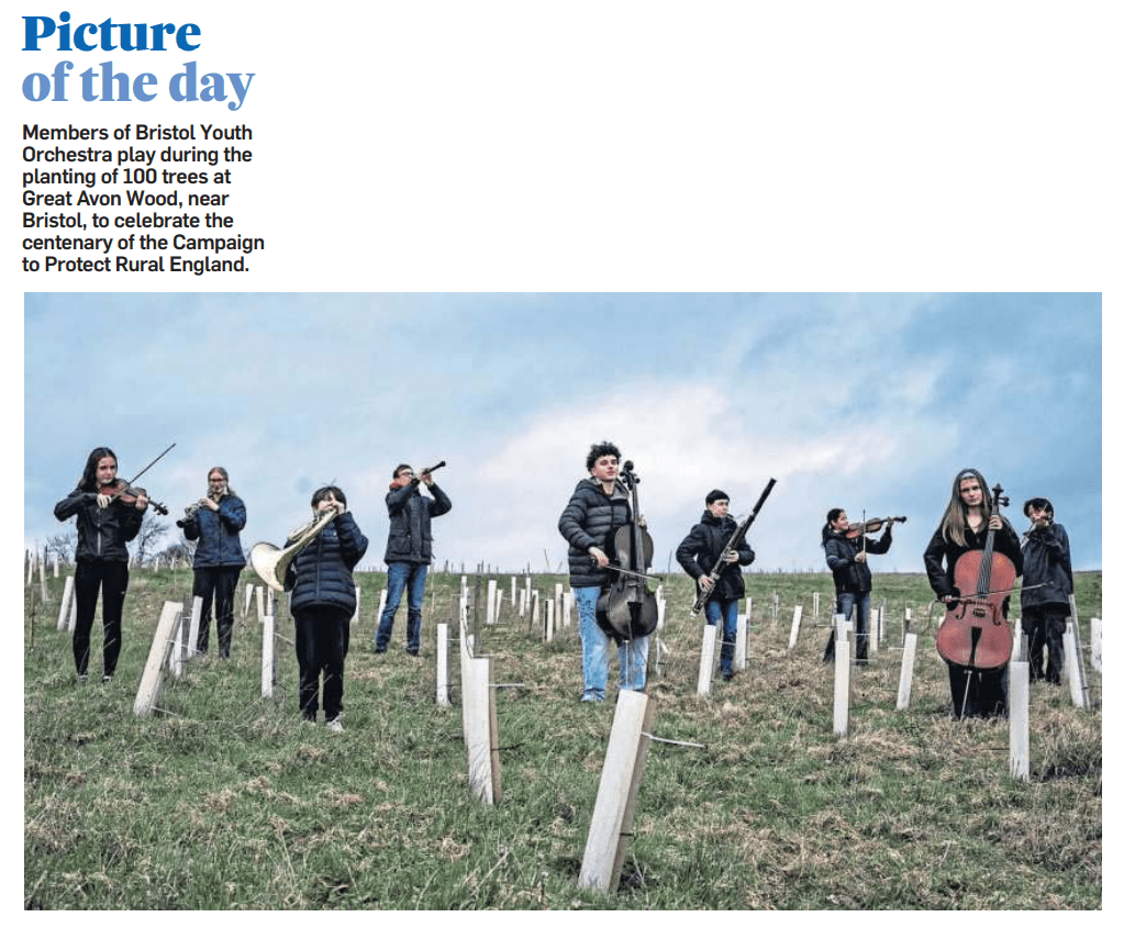 A photo of 10 young musicians standing amongst a newly-planted copse of trees, holding a mix of orchestral instruments