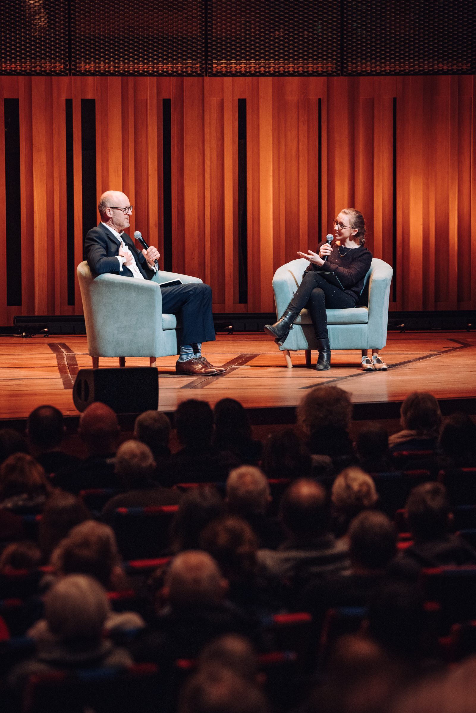 A man in a suit and glasses sits on stage in an arm chair as he interviews a female musician with long hair in a ponytail also sitting in an armchair.