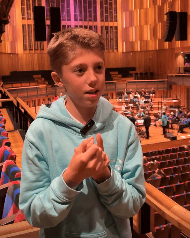 A young person wearing a light blue school jumper is interviewed on a balcony in a large concert hall.