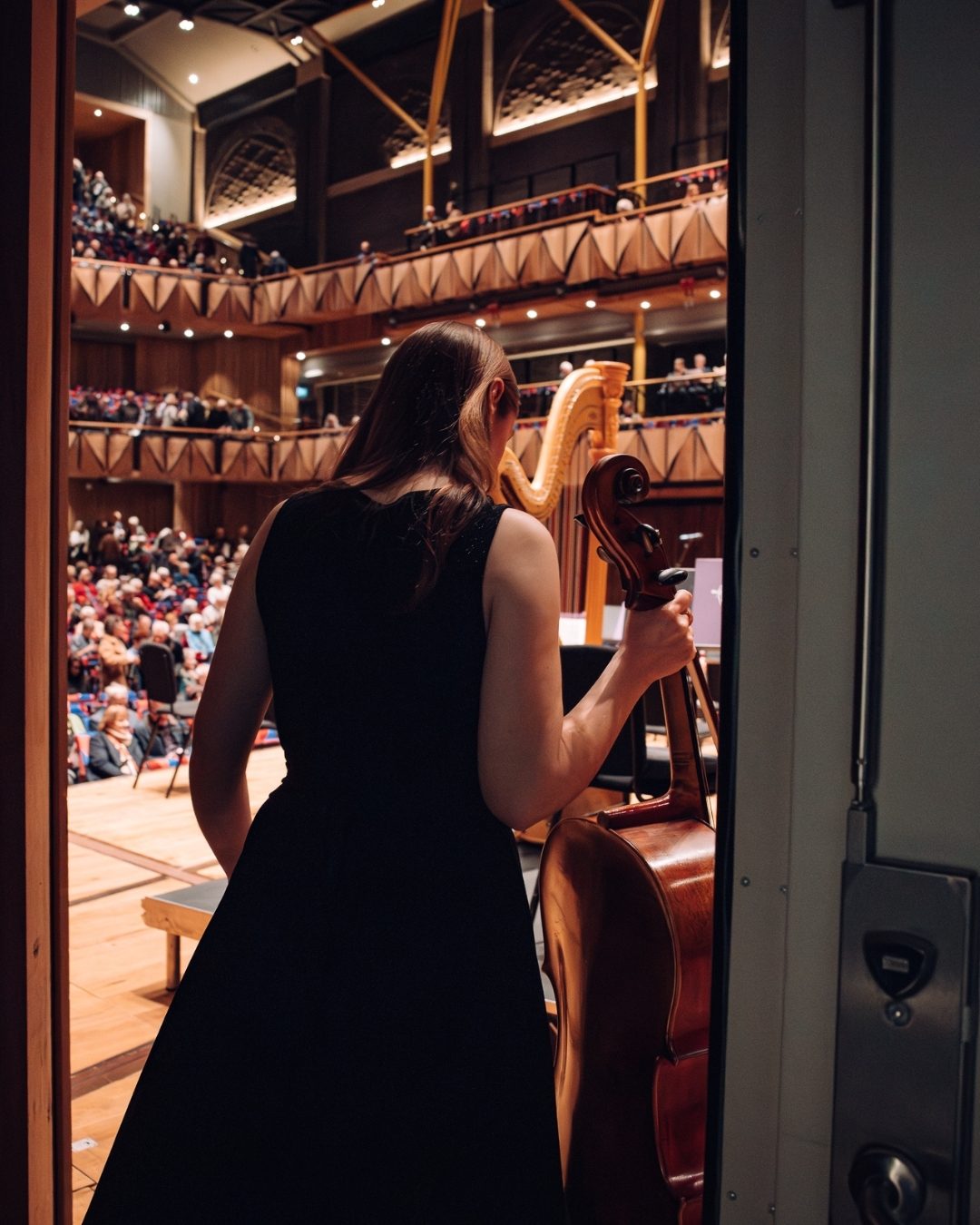A cellist dressed in black walks through a stage door holding their instrument onto a large stage with an orchestra in an auditorium full of people.