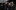 Seven people pose together, all dressed in black in front of a black, metallic backdrop.