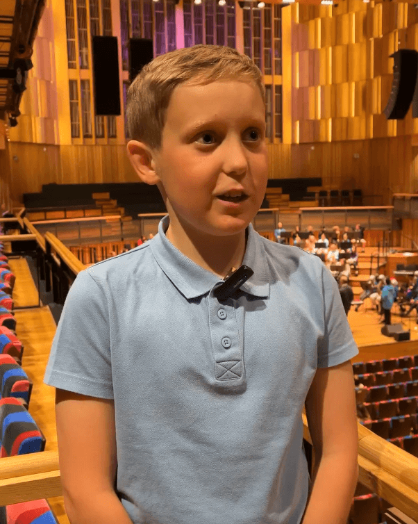 A young person wearing a light blue school shirt is interviewed on a balcony in a large concert hall.