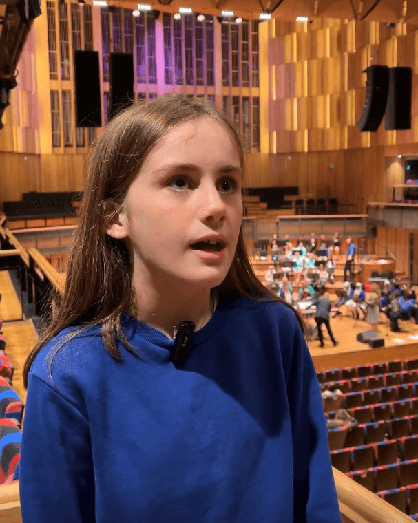 A young person wearing a blue school jumper is interviewed on a balcony in a large concert hall.