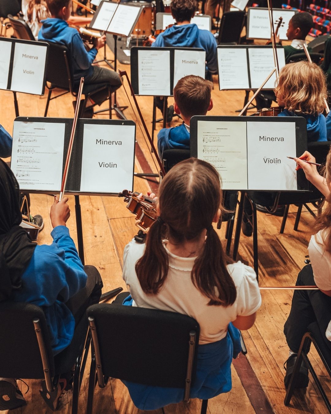 School children rehearse on stage as they sit in an orchestra with sheet music