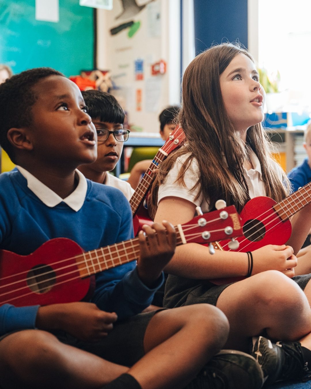 School children sit cross legged playing ukulele looking delighted