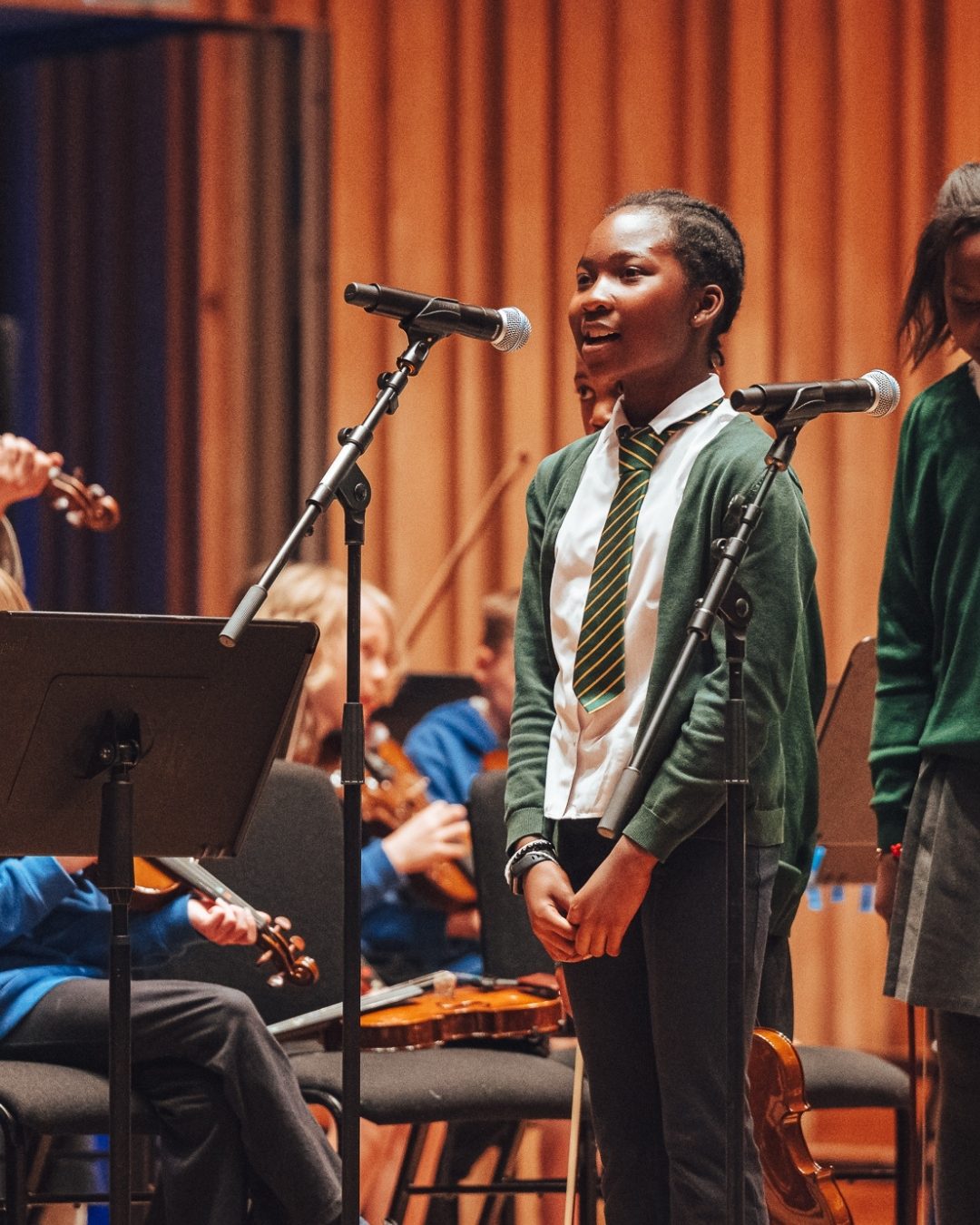A young person wearing a green school jumper sings into a microphone on stage