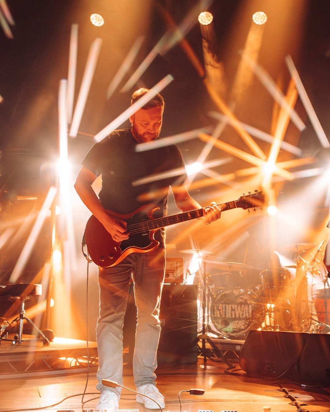 A guitarist, bassist and a keyboard player all perform live on stage at a concert hall, engulfed in bright orange floodlights.