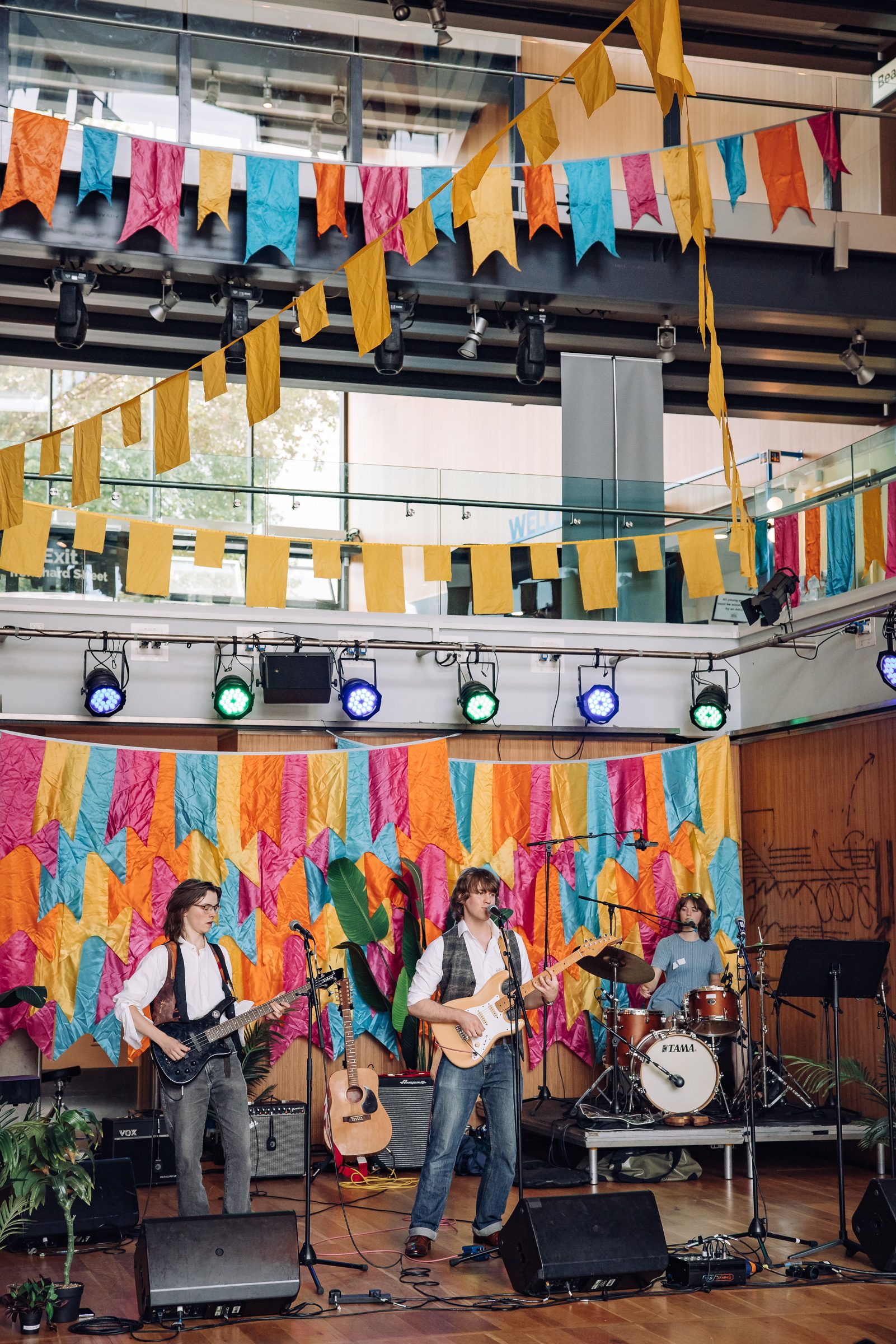 Three musicians perform on stage with guitars and drums under multicolored flags and stage lights in an indoor venue.