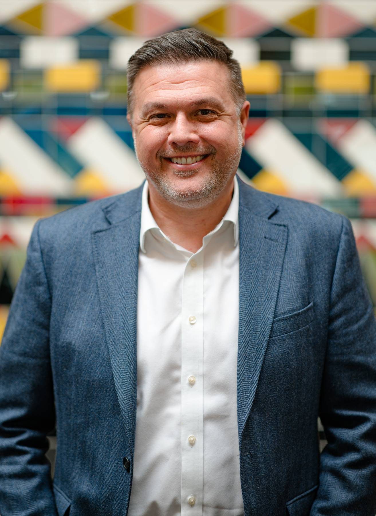 A smiling person wearing a blue suit jacket over a white button-up. Behind them is a tiled wall consisting of multi-coloured geometric shapes.
