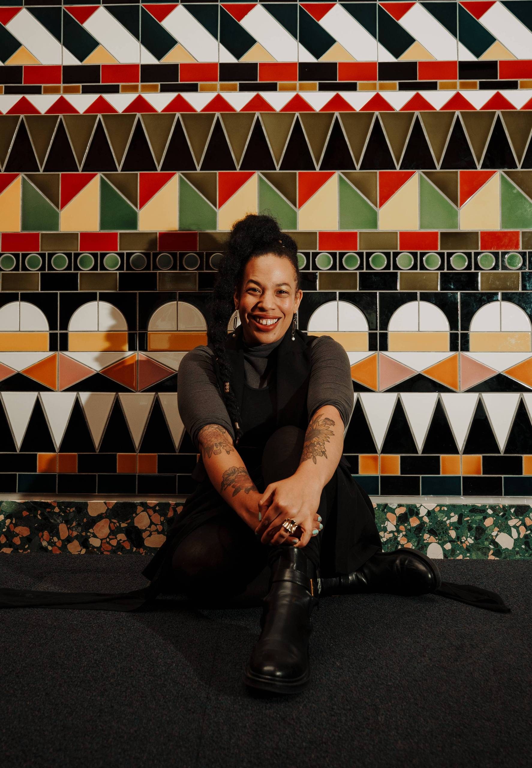 A smiling person with long dark hair tied up sits in front of a tiled wall made up of multi-coloured geometric shapes. They pose with their hands clasped together over an outstretched knee.