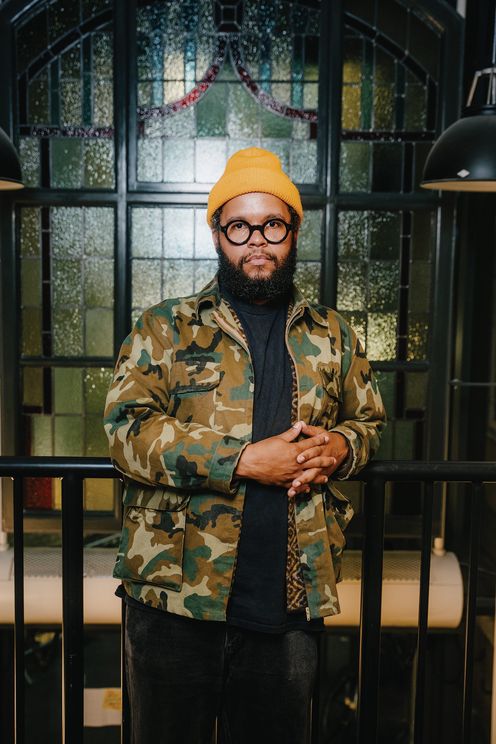 A person wearing a camouflage print jacket, yellow beanie and black, thick-rimmed glasses leans on a black railing in front of a stained-glass window.