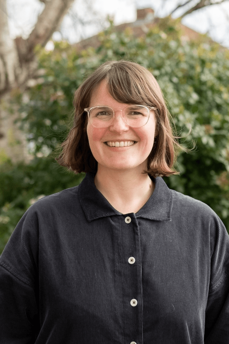 A person with shoulder length brown hair wearing large transparent glasses stands outside and smiles