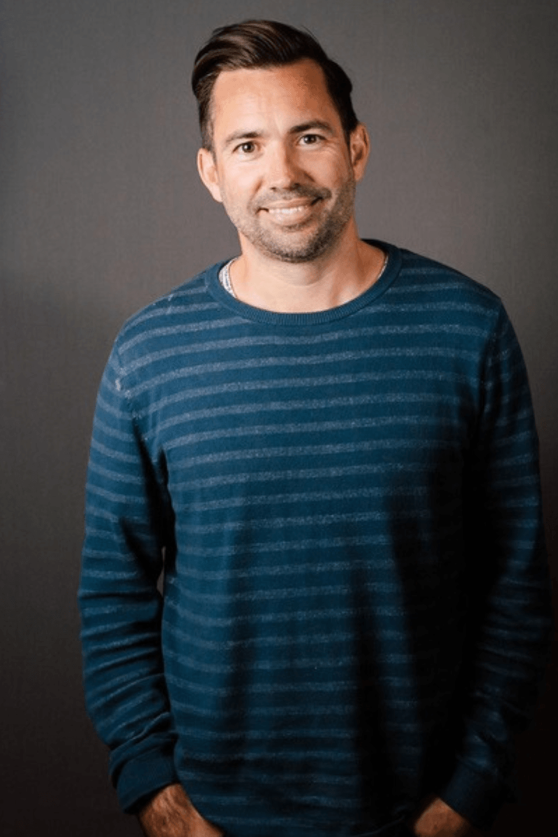 A person with short brown hair wearing a dark blue striped shirt smiles